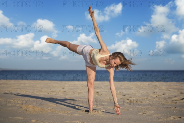 A woman practices Ardha Chandrasana Half Moon Pose on sandy beach while balancing on one leg and reaching to the ground