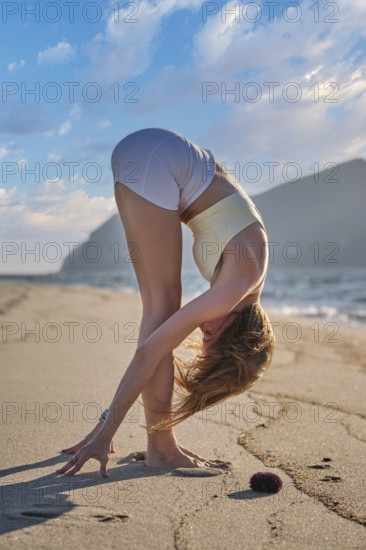 A woman performs Uttanasana, a forward bend yoga pose, on the beach as the sun sets over the ocean, creating a beautiful view