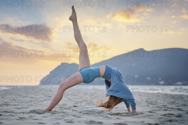 A woman performs Eka Pada Urdhva Dhanurasana on a sandy beach as the sun sets in the background, showcasing her yoga practice