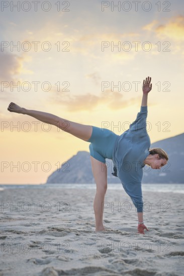 A woman does Ardha Chandrasana Half Moon Pose on the sand during sunset. She balances on one leg while her other leg is extended
