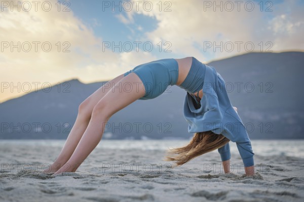 A woman practices Urdhva Dhanurasana on a sandy beach while the sun sets behind mountains. The sky is filled with warm colors