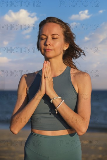 A woman practices yoga on the beach at sunset, holding hands in Namaskar Mudra as she meditates with her eyes closed