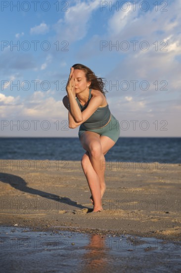 A woman practices standing Garudasana on a beach with one leg wrapped around the other at sunset by the water