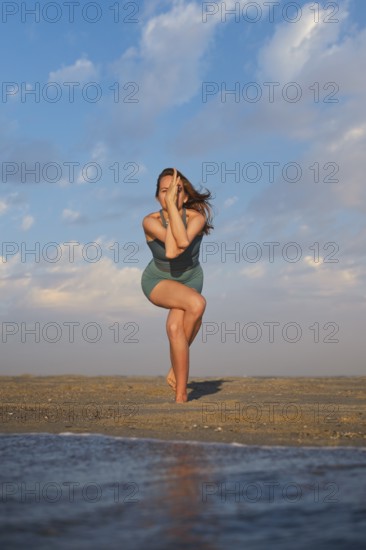 A woman stands on a sandy beach performing Eagle Pose with wrapped legs and crossed arms during sunset, showcasing balance and strength