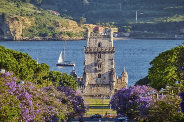 Blooming jacaranda trees line the street as Belem Tower stands majestic over the Tagus River at sunset with tourist sailboats nearby
