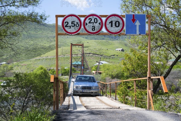 A car crosses a suspension bridge, accompanied by road signs with width and height restrictions over green hills, Kura River near Nijgori, Samtskhe-Javakheti region, Samtskhe-Javakheti, Georgia