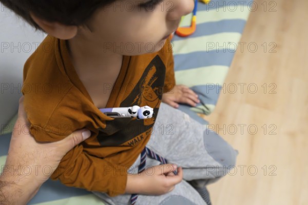 Child wearing a t shirt having a digital thermometer placed under arm for temperature measurement, indicating illness, flu, or a fever, with a parents arm providing assistance and comfort