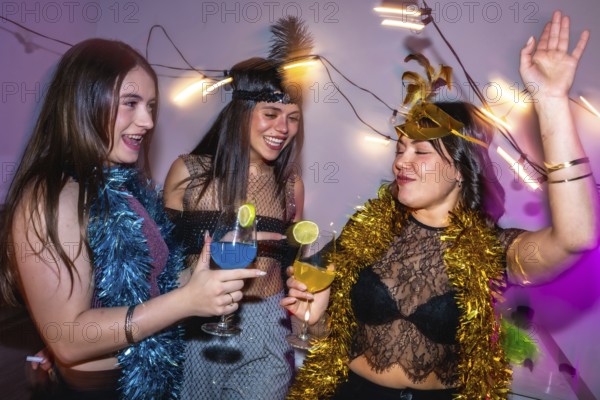 Three young women smiling, laughing, and drinking colorful cocktails while having fun at a festive new year's eve or costume party, surrounded by sparkling string lights and tinsel decorations