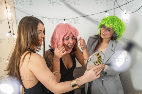 Group of happy young women friends trying on fun colorful wigs and preparing for a costume party or new year's eve celebration at home, sharing moments of joy and friendship