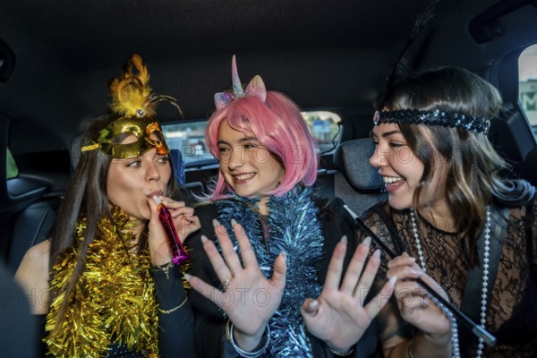 Three young women in festive costumes ride in a car, wearing tinsel and makeup, laughing and blowing party blowers as they celebrate a joyful night out together