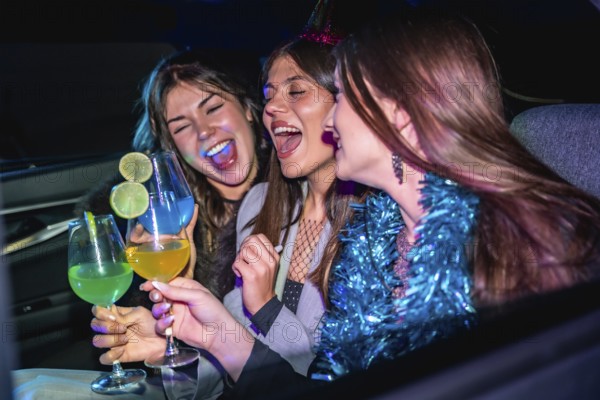 Three young women happily celebrating and laughing together in a car at night, holding colorful cocktails and enjoying a fun festive moment with friends