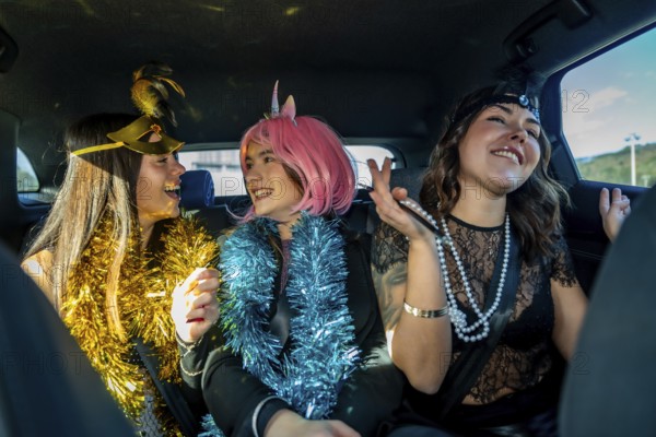 Three young women wearing festive costumes and tinsel garlands, smiling and laughing together while traveling in the back seat of a car, enjoying a happy celebration