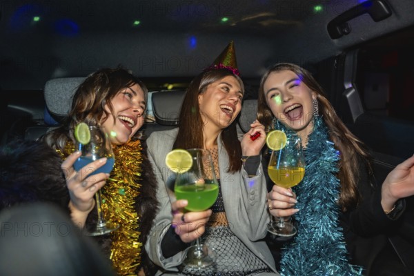 Three friends dressed up in party hats and tinsel garlands laughing and toasting colorful cocktails inside a car, enjoying a festive night out full of joy and spontaneous fun