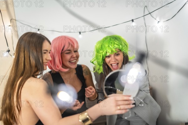 Two young women in pink and green wigs laugh and celebrate at a colorful house party, sharing joyful candid moments, friendship and playful festive fun indoors with lights and decorations