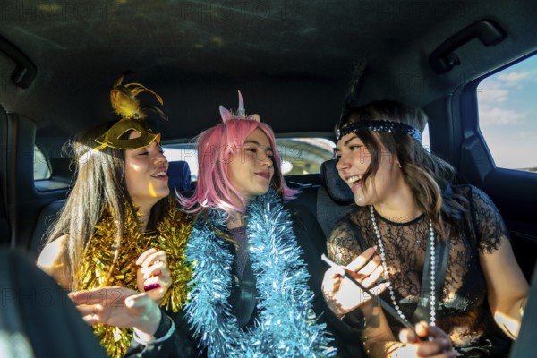 Three young women laugh and celebrate in a car during a colorful costume party, wearing masks, wigs and tinsel for a festive, carefree road trip or holiday celebration