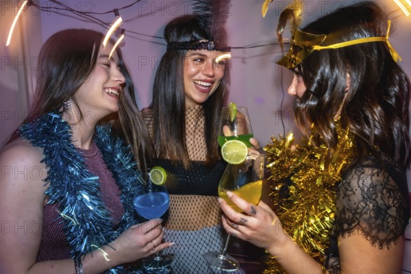 Young women friends laughing and cheering with colorful cocktails, celebrating together during a festive new year's eve or costume party with tinsel decorations
