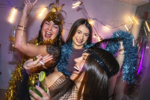 Three young women laughing and dancing, celebrating a festive new year's eve or costume party at home, wearing fun accessories and drinking cocktails in a lively atmosphere