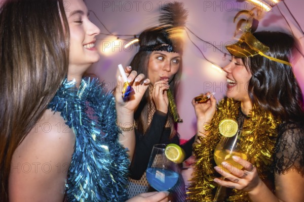 Three happy young women enjoying a lively new year's eve or costume party, wearing festive accessories, blowing party horns, and holding cocktails with bright, colorful drinks