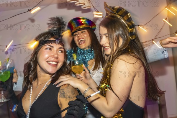 Young women in festive costumes laugh and toast cocktails at a colorful new year's eve home party, sharing joyful friendship, confetti, and lively celebration together