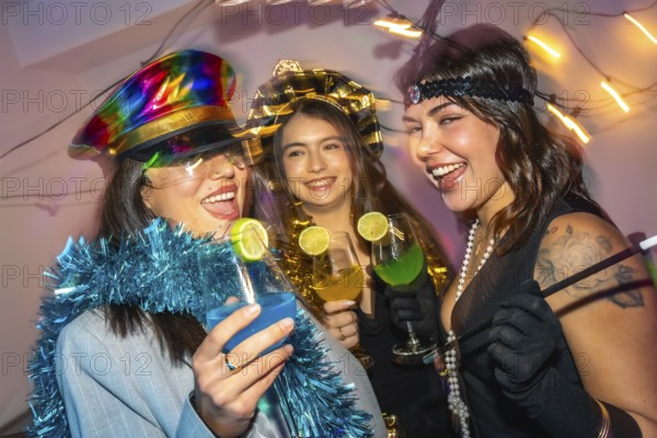 Three happy young women laughing and drinking colorful cocktails while celebrating a new year's eve or costume party with festive decorations and glowing string lights