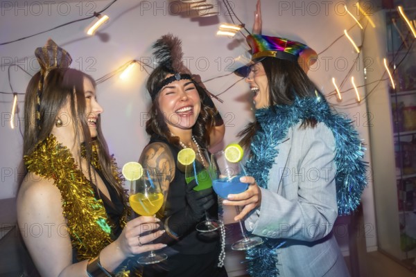 Three diverse young women friends wearing festive costumes and tinsel, celebrating a new year's eve or costume party at home, laughing and toasting with colorful cocktails in wine glasses