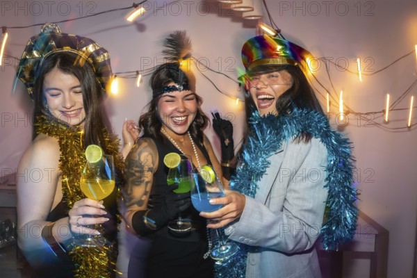 Three young women friends enjoying a fun new year's eve party, celebrating with colorful cocktails, dressing up in costume hats and festive tinsel while laughing together