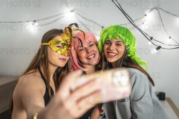 Group of happy young women celebrating new year's eve or a costume party indoors, smiling and posing for a cheerful selfie while wearing colorful wigs and a masquerade mask