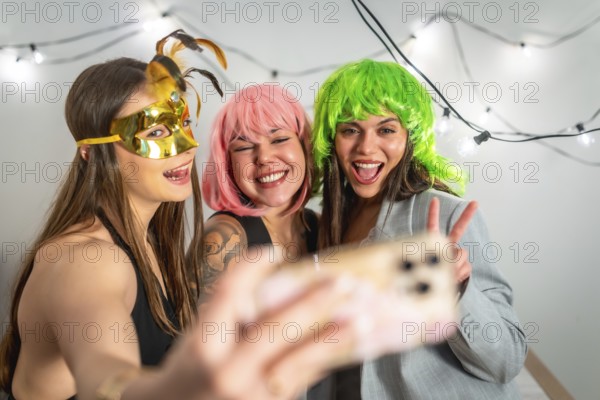 Girls friends celebrating a new year's eve or costume party, laughing and taking a selfie with a smartphone, wearing colorful wigs and a golden mask, lights decorating the background