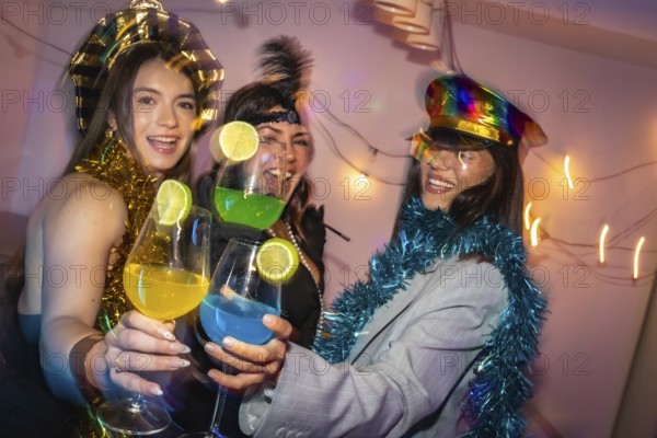 Three happy young women celebrating a party, toasting colorful cocktails with lime slices, laughing and having fun with festive decorations and costumes in a vibrant home setting
