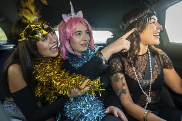 Three young women dressed in cheerful costumes with tinsel on the back seats of a car, joyfully interacting and laughing while pointing at something outside