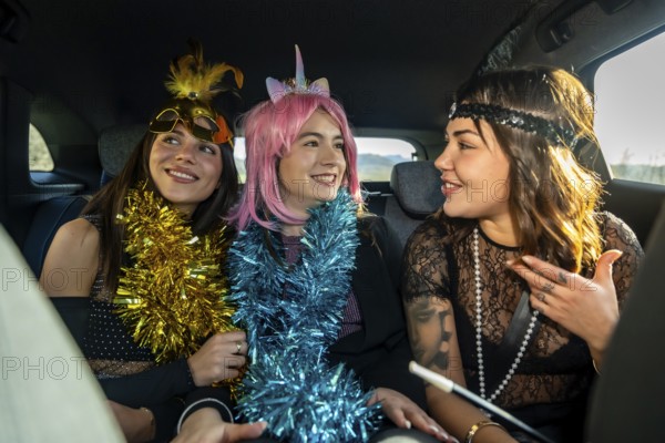 Three young women in colorful costume accessories and party attire laughing and talking in the back seat of a car, celebrating a special event together
