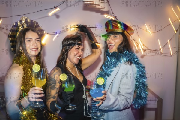 Three diverse women wearing festive costumes, decorations, and holding colorful cocktails, enjoying a lively friends costume party at home, celebrating an event