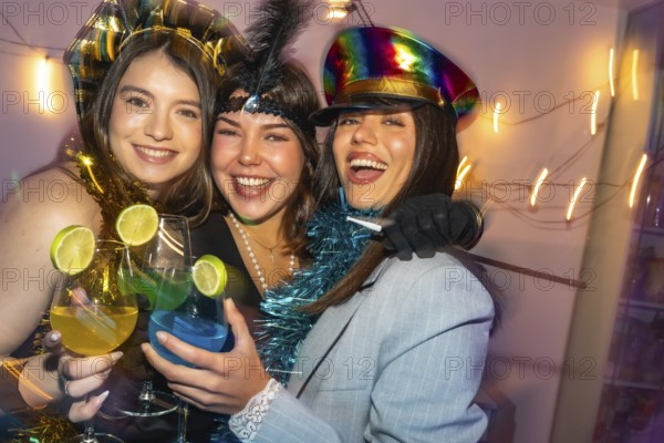 Three happy young women laughing and toasting with colorful cocktails, dressed in costumes, celebrating a festive gathering with friends indoors in a vibrant and lively atmosphere