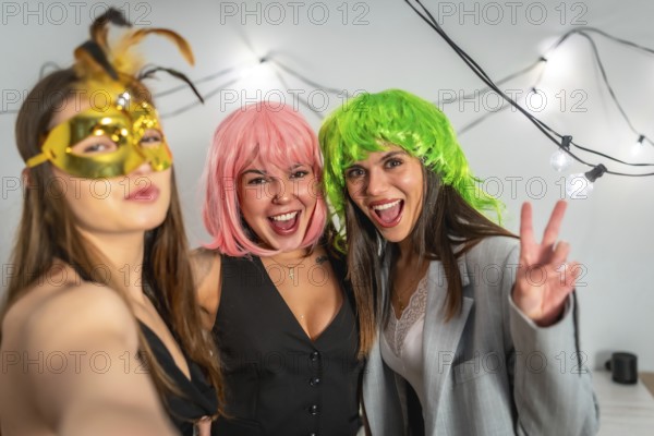 Three young women in colorful wigs and a masquerade mask laugh and pose for a bright, carefree selfie at a festive costume party with string lights in the background