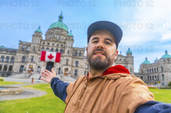 Happy bearded tourist taking a selfie with outstretched arms in front of the parliament buildings with a canadian flag hanging from the facade in victoria, british columbia, canada