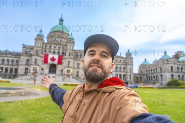 Happy bearded tourist taking a selfie with outstretched arm in front of the parliament buildings in victoria, british columbia, with canadian flag displayed prominently