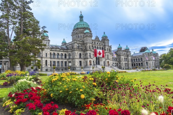 Canadian flag waving in front of parliament buildings in victoria, british columbia, canada during summer with colorful flowers blooming in the foreground and a bright blue sky