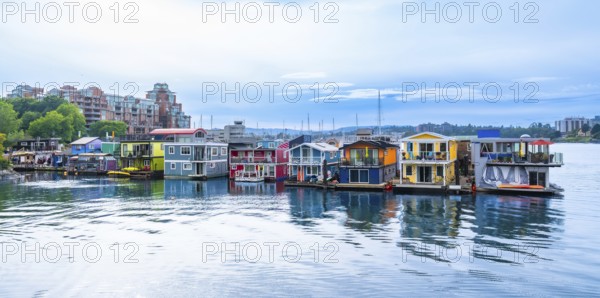 Colorful floating homes at fisherman's wharf park are reflecting in the calm water in victoria, british columbia, canada, with modern buildings and cloudy sky in the background during a summer day