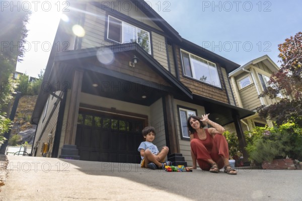 Mother and son are sitting on their driveway in front of their house, enjoying quality time together on a bright summer day, waving and playing with toy cars