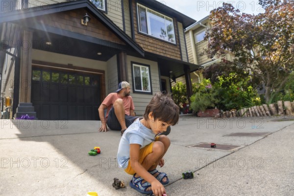 Young boy plays with toy cars on the driveway of a modern home while his father watches from behind, creating a heartwarming scene of family bonding and childhood joy