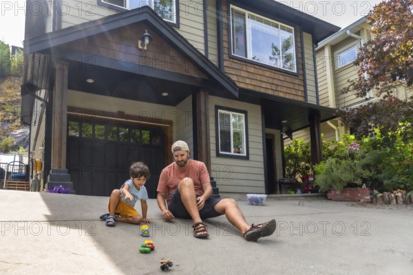 Father and son are sitting on their driveway, playing with a collection of toy cars in front of their suburban home on a sunny summer day