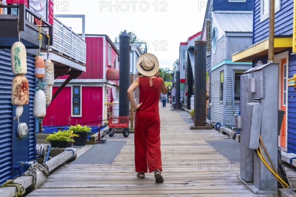 Tourist wearing a straw hat and a red jumpsuit is walking on a wooden boardwalk between colorful floating houses in fisherman's wharf park in victoria, british columbia, canada