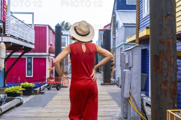 Tourist wearing a red jumpsuit and straw hat walking at fisherman's wharf park, a colorful floating house community, in victoria, british columbia, canada