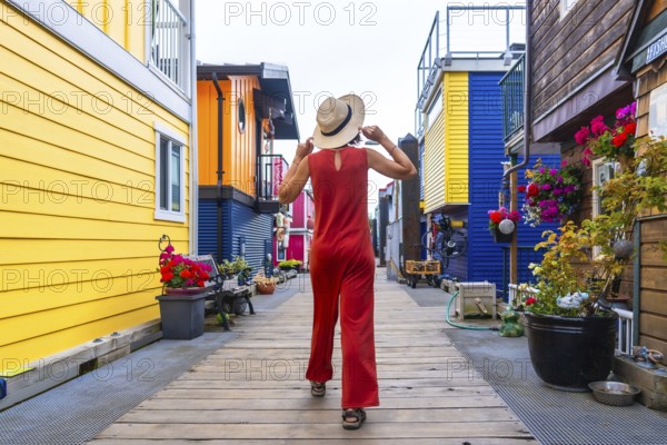 Tourist wearing a straw hat and red jumpsuit walking along a wooden walkway by vibrant floating houses at fisherman's wharf park in victoria, british columbia