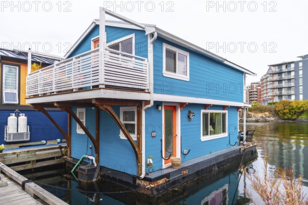 Colorful floating home with a white balcony and orange door reflecting in calm waters of victoria harbor, surrounded by condos on a cloudy day