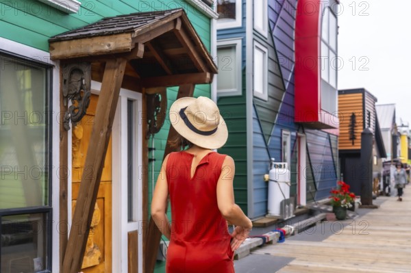 Female tourist wearing a straw hat and a red dress is enjoying a summer day walking down the fisherman's wharf park in victoria, british columbia, canada, admiring the colorful floating houses
