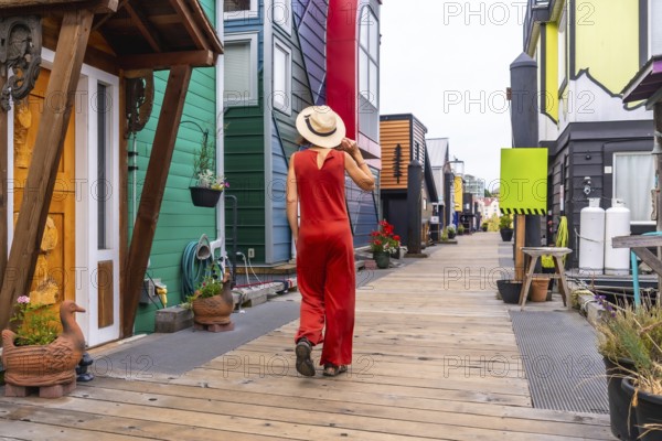 Tourist wearing a straw hat and a red jumpsuit is walking on a wooden walkway exploring the colorful floating homes of fisherman's wharf park in victoria, british columbia, canada