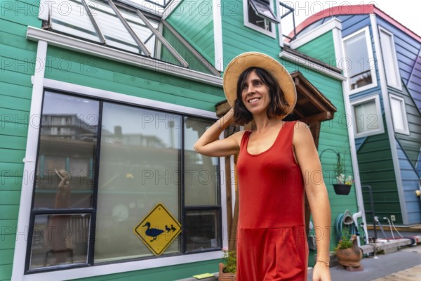 Happy tourist adjusting her straw hat while walking and smiling in front of colorful floating houses in fisherman's wharf park in victoria, british columbia