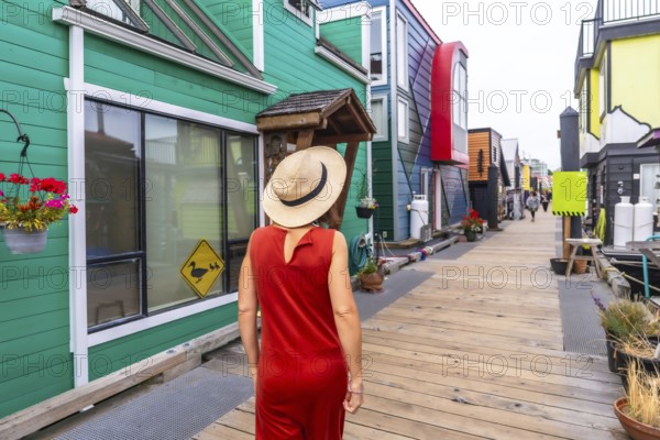 Tourist wearing a straw hat and orange dress walking along a wooden walkway, visiting vibrant floating houses at fisherman's wharf park in victoria, british columbia on a sunny summer day