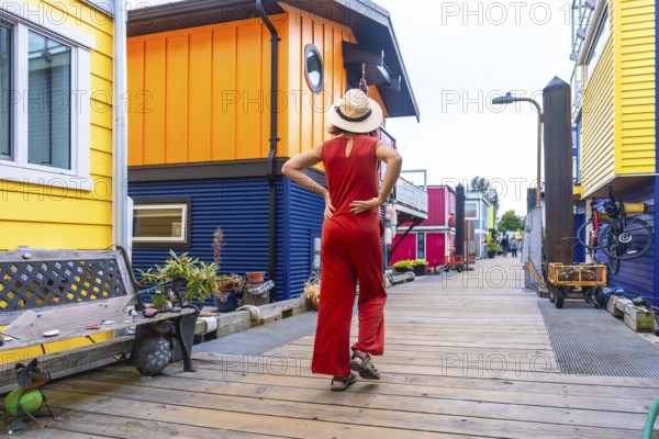 Young woman tourist in a straw hat and red jumpsuit walking along a wooden boardwalk, admiring colorful floating houses at fisherman's wharf park in victoria, british columbia